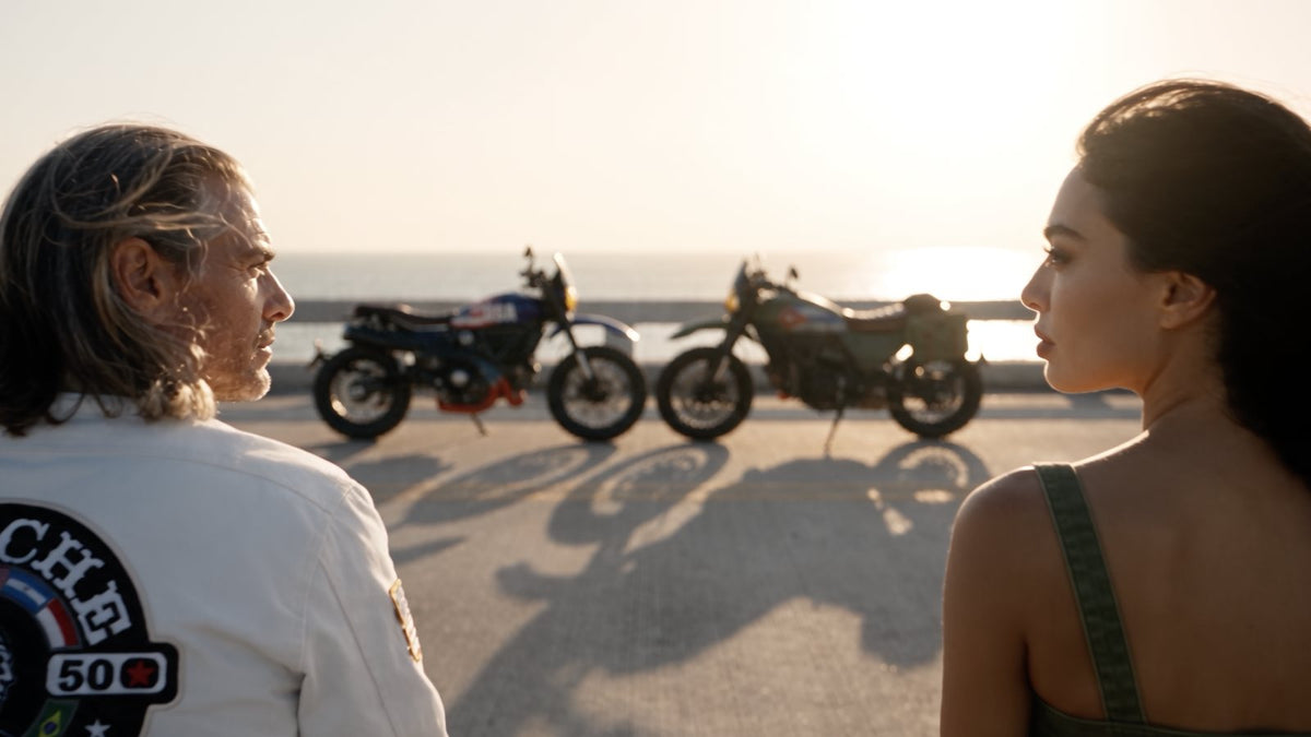 A man in a white APACHE STAR jacket and a woman face each other on the bridge at sunset, the blue and the olive-green motorcycle parked behind them, in the film THE BRIDGE: 90 miles apart