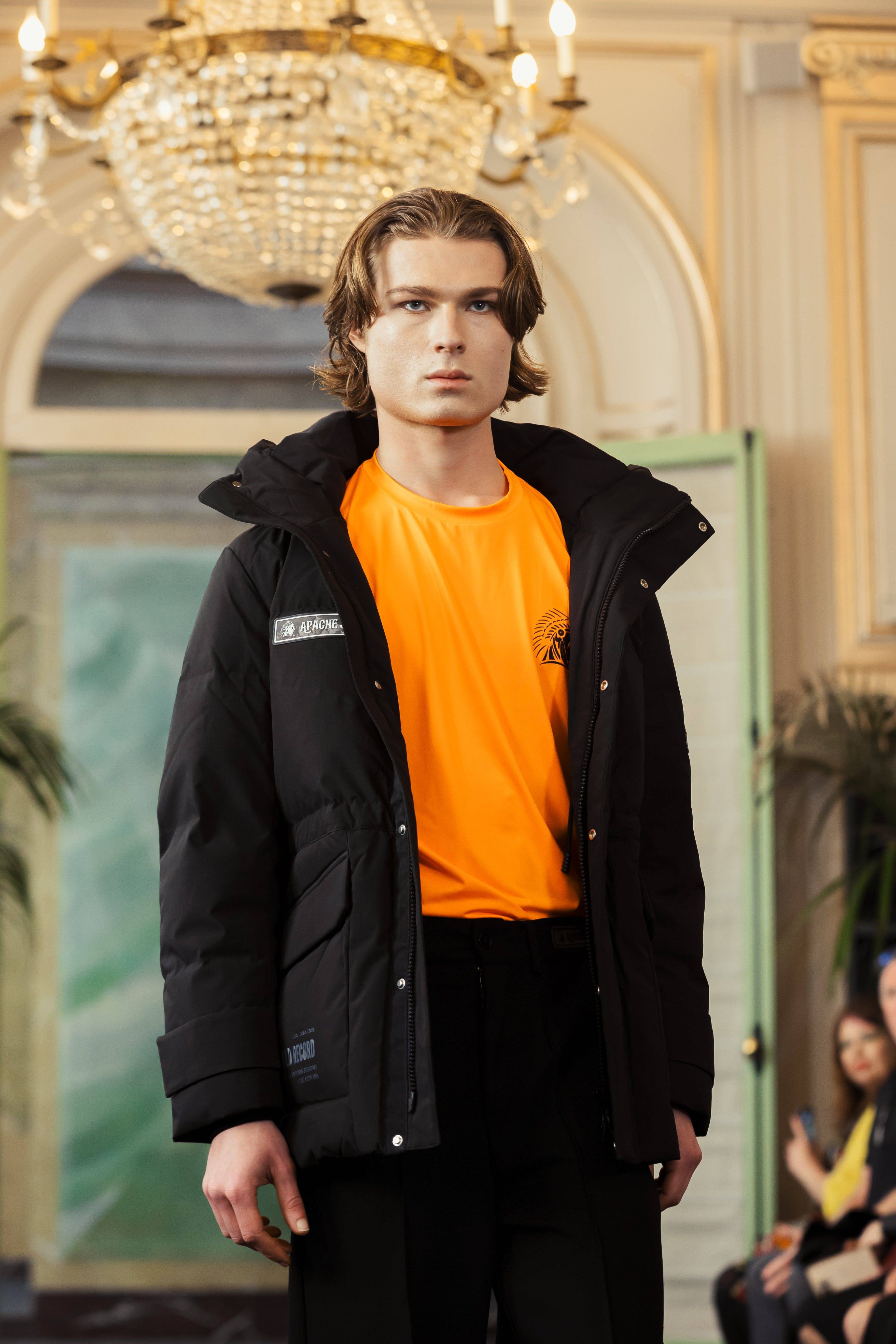 Runway close-up at Paris Fashion Week 2025 of APACHE STAR BURNING BATTALION Longsleeve Shirt styled under the black AMERICAN ANCHORAGE down jacket, highlighting its vibrant color and chest logo.