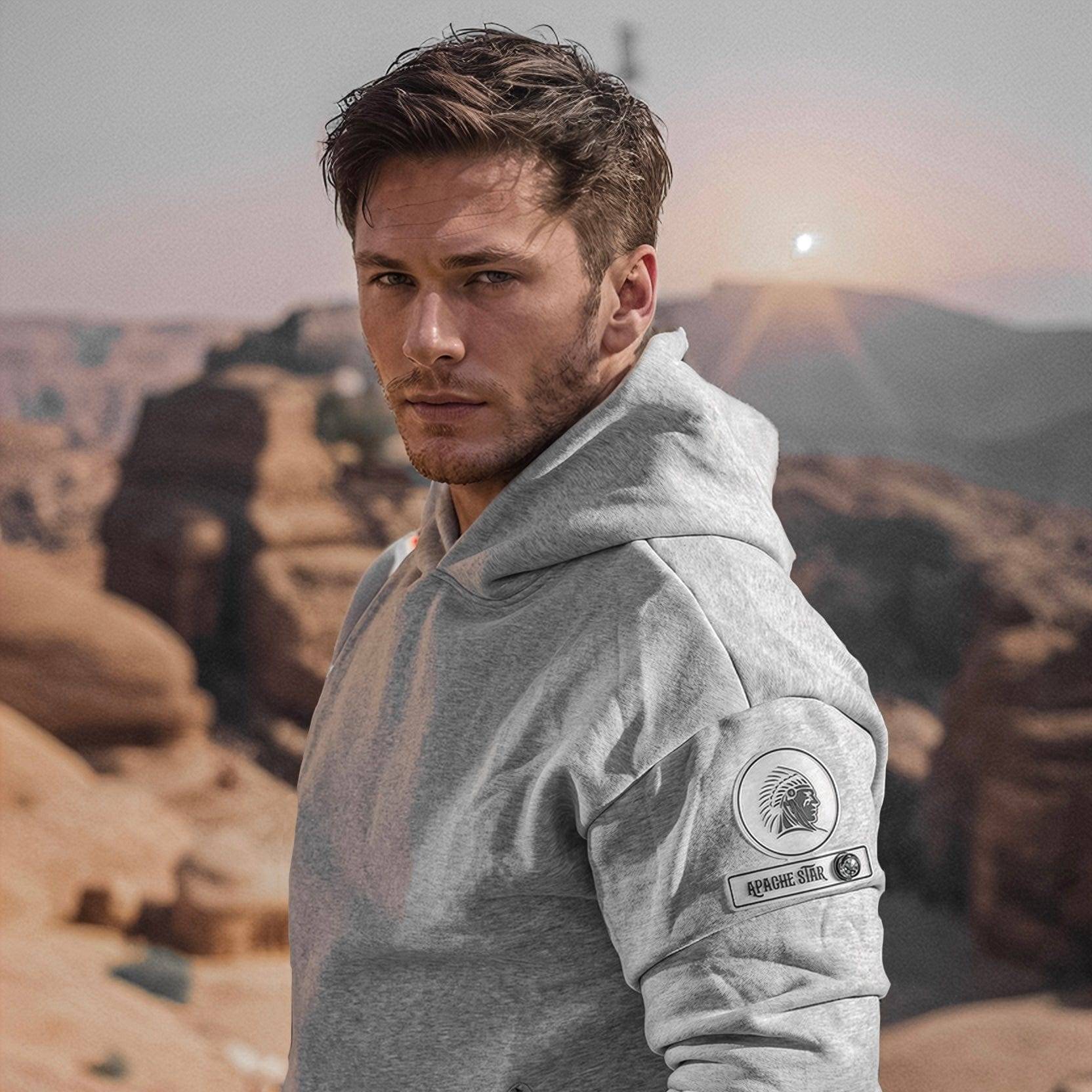 Man in grey Iconic Dubai Hoodie by APACHE STAR standing in a desert landscape. Showcases luxury compass patch and rugged outdoor appeal.