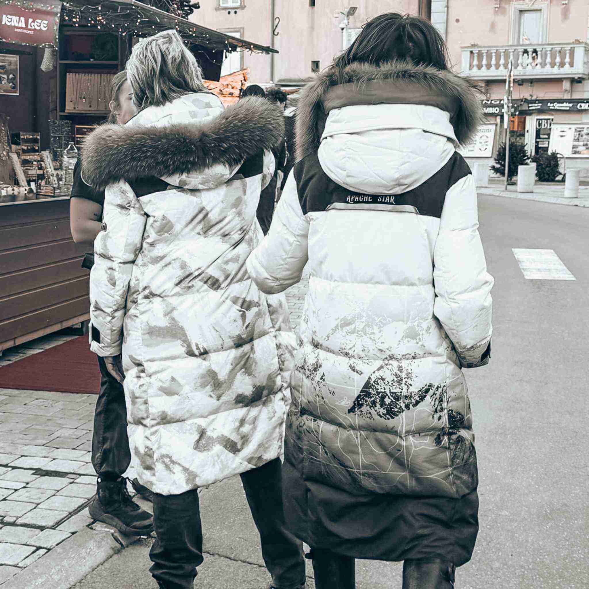 Back view of two women in Trident Trooper Down Coats by APACHE STAR with fur hoods in St. Tropez, highlighting the coat’s mountain print and winter appeal.