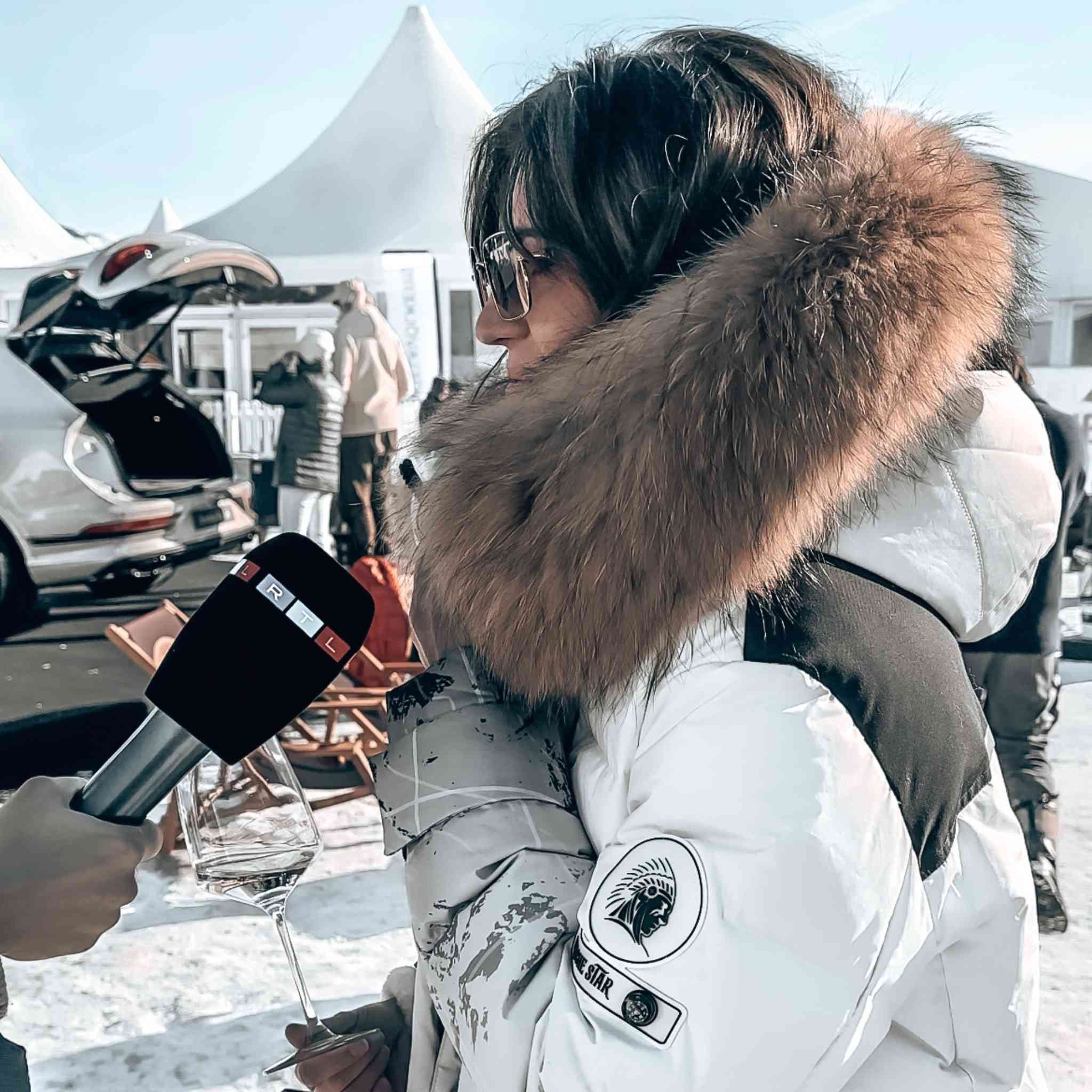 Person in white winter jacket with fur hood holding a glass of champagne at snowy outdoor event