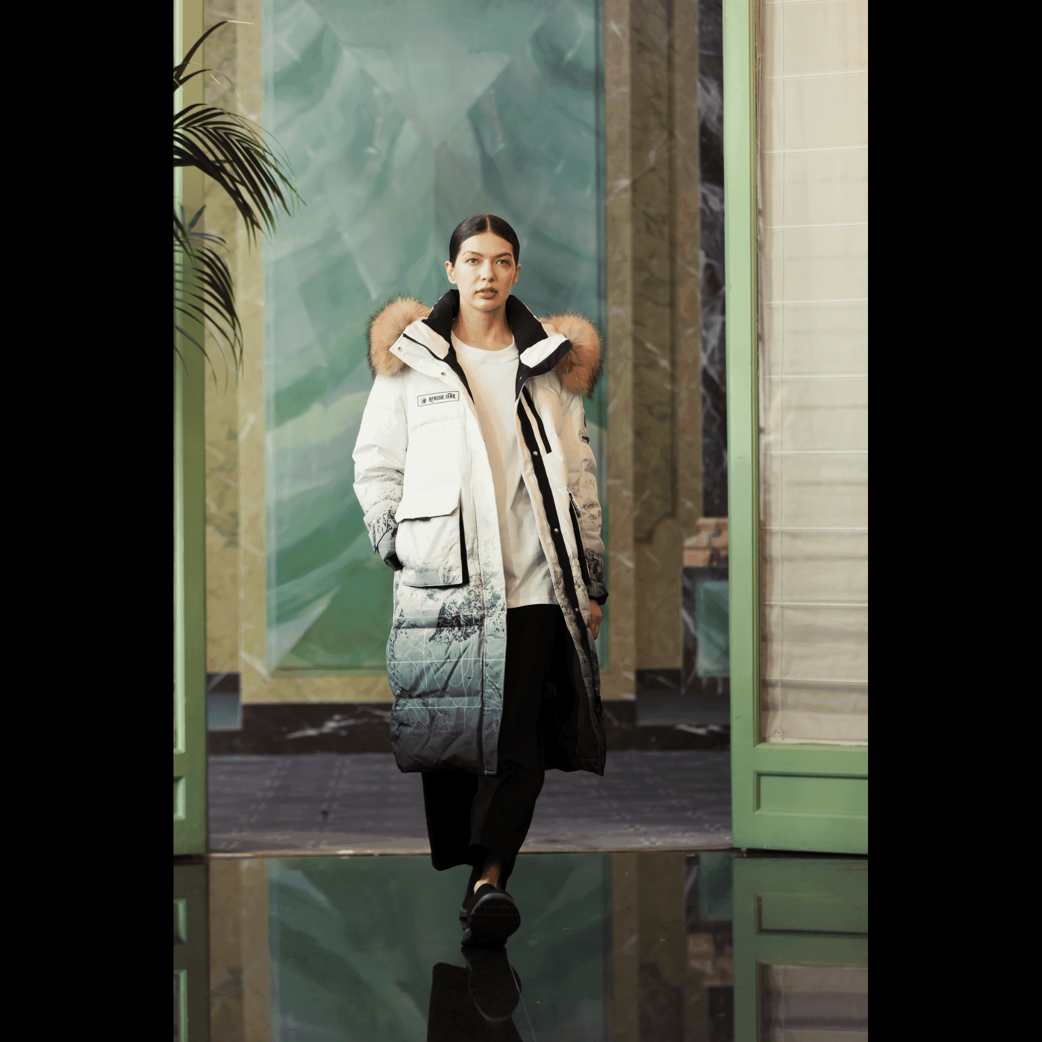 Model walking indoors wearing Trident Trooper Down Coat by APACHE STAR, showcasing stylish winter fashion with mountain print and fur hood at Paris Fashion Week.