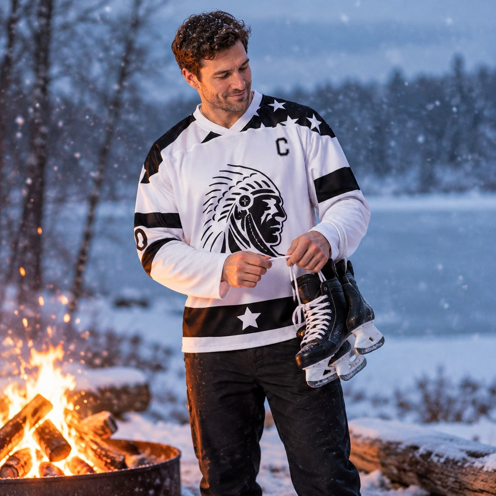 A man lacing ice skates beside a campfire in falling snow, wearing the white APACHE STAR ice hockey jersey "Frozen Fifty"