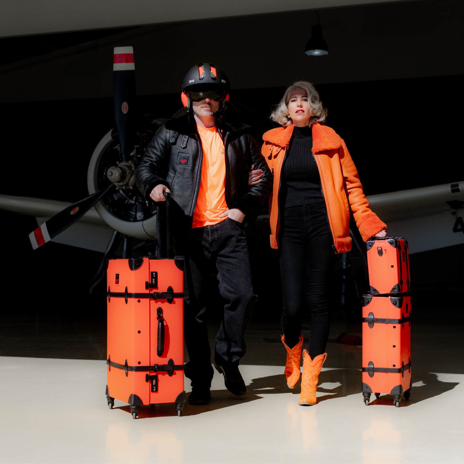 Fashionable travelers with APACHE STAR® Heritage Trunk Set in Apache Orange, showcasing luxury suitcase travel set in an aviation-hangar.